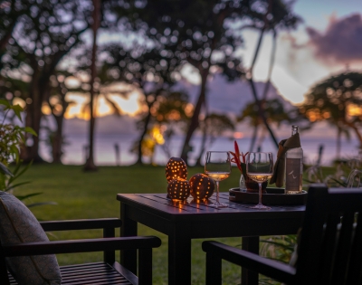 A cozy outdoor setting with two glasses of wine, a bottle, and illuminated decor, surrounded by lush trees at sunset.