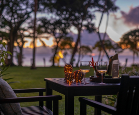 A cozy outdoor setting with two glasses of wine, a bottle, and illuminated decor, surrounded by lush trees at sunset.