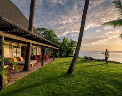 A tranquil beachfront villa with palm trees, green lawn, and a person enjoying the sunset by the ocean.