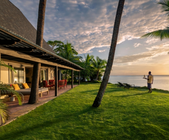 A tranquil beachfront villa with palm trees, green lawn, and a person enjoying the sunset by the ocean.