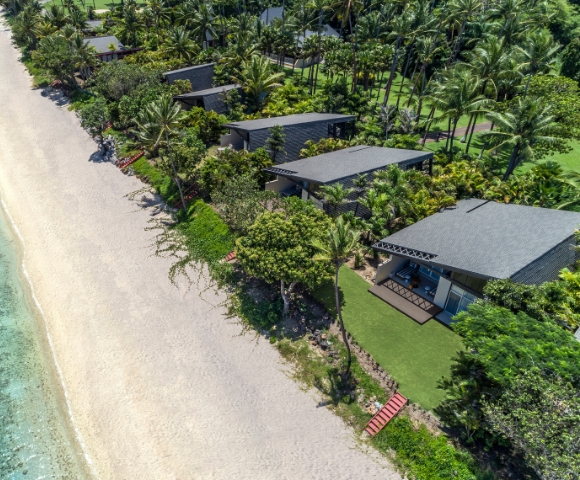 Aerial view of tropical beach with palm trees and modern beachfront bungalows along the shore. Clear water and sandy beach visible.
