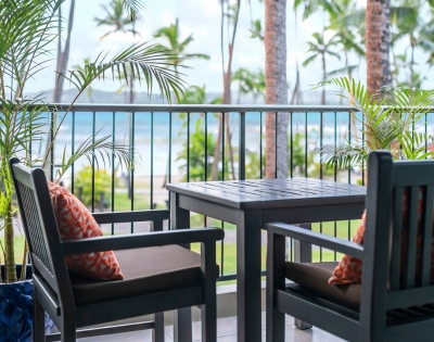 Cozy balcony with two chairs and a table overlooking the beach, framed by tropical plants and palm trees.