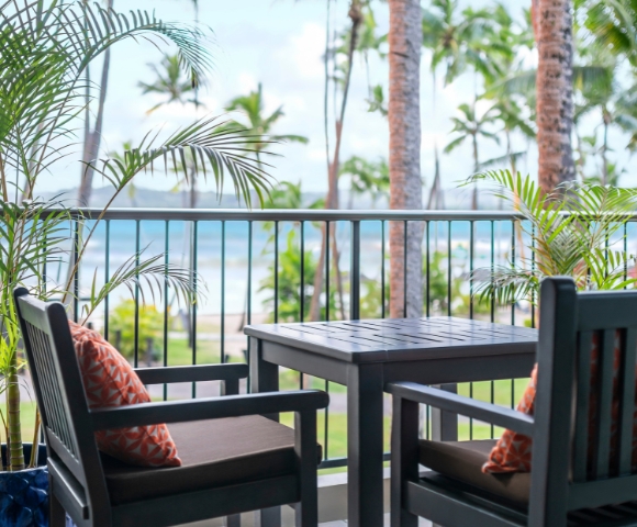 Cozy balcony with two chairs and a table overlooking the beach, framed by tropical plants and palm trees.
