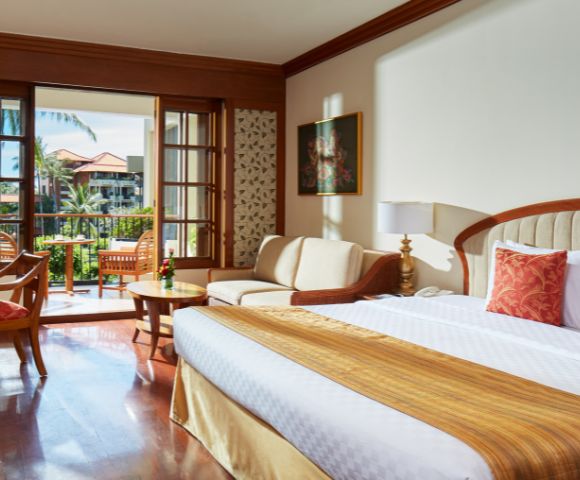 A spacious hotel room with a king-sized bed, seating area, and balcony overlooking lush greenery and palm trees. Bright and inviting.