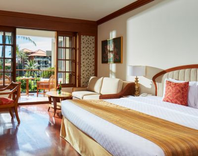 A spacious hotel room with a king-sized bed, seating area, and balcony overlooking lush greenery and palm trees. Bright and inviting.