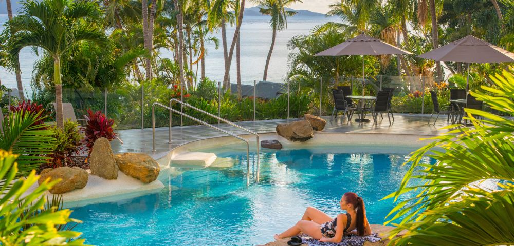 A woman relaxes on a rock by a serene pool surrounded by palm trees, with beach umbrellas in the background and ocean views.