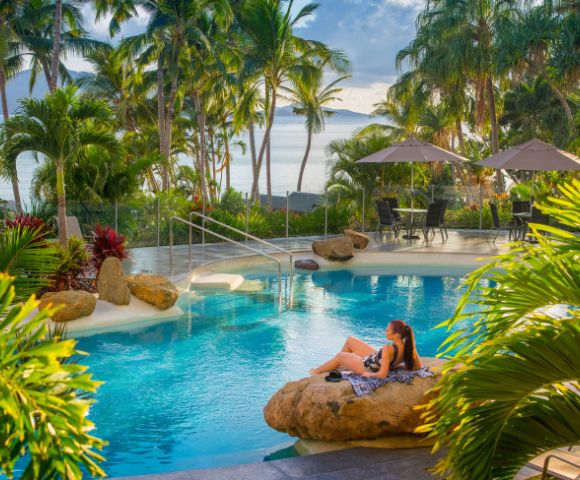 A woman relaxes on a rock by a serene pool surrounded by palm trees, with beach umbrellas in the background and ocean views.
