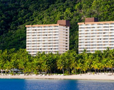 Two beachfront hotel towers surrounded by lush tropical trees and clear blue water, set against a green hillside.