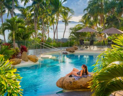 A woman relaxes on a rock by a serene pool surrounded by palm trees, with beach umbrellas in the background and ocean views.
