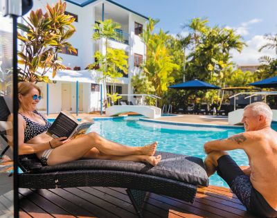 Two people relax by a sunny poolside, one reading a book on a lounge chair, surrounded by lush tropical greenery and blue umbrellas.