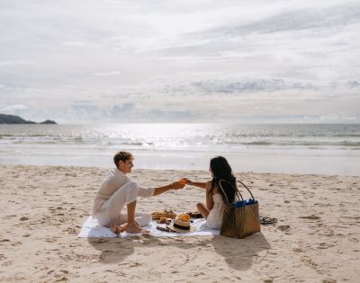 A couple enjoys a picnic on the beach, sharing fruit under a bright sky with ocean waves in the background.