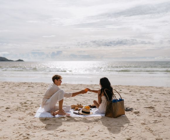 A couple enjoys a picnic on the beach, sharing fruit under a bright sky with ocean waves in the background.