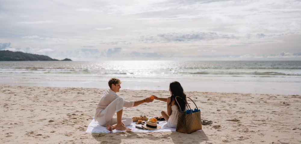A couple enjoys a picnic on the beach, sharing fruit under a bright sky with ocean waves in the background.