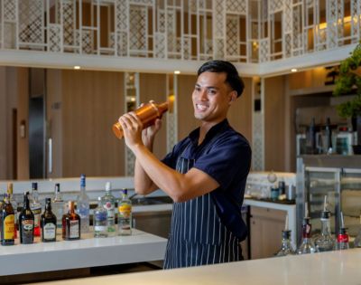 A bartender in a striped apron skillfully shakes a cocktail shaker, surrounded by an array of liquor bottles on the bar.