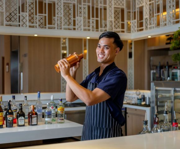 A bartender in a striped apron skillfully shakes a cocktail shaker, surrounded by an array of liquor bottles on the bar.