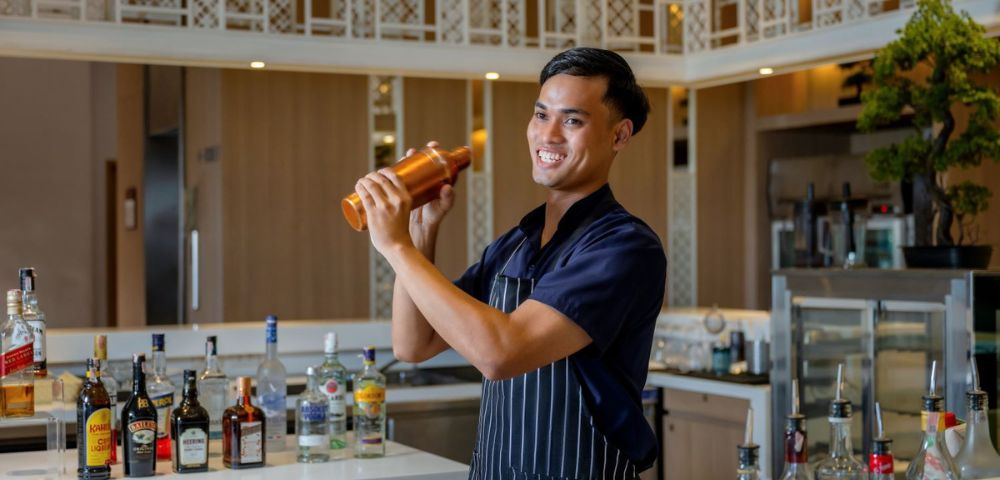 A bartender in a striped apron skillfully shakes a cocktail shaker, surrounded by an array of liquor bottles on the bar.