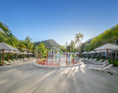 Colorful water play area with fountains, surrounded by lounge chairs and palm trees, set against a mountain backdrop and clear blue sky.