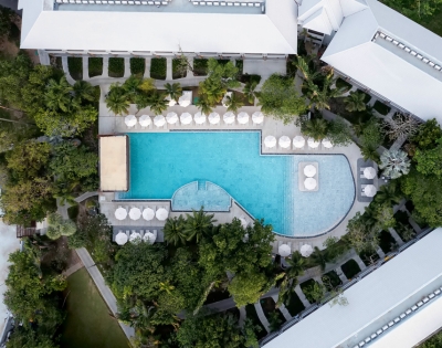Aerial view of a resort pool, surrounded by lush greenery and lounge chairs, with modern buildings in the background.