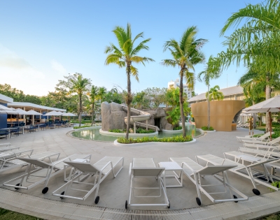 A serene pool area featuring lounge chairs, a rock slide, and lush palm trees, set against a clear blue sky. Ideal for relaxation.