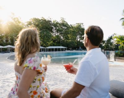 A couple enjoys tropical cocktails by a serene poolside at sunset, surrounded by lush greenery and white cabanas.