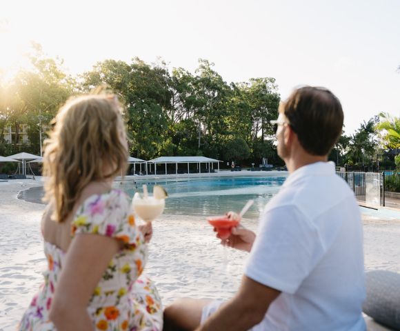 A couple enjoys tropical cocktails by a serene poolside at sunset, surrounded by lush greenery and white cabanas.