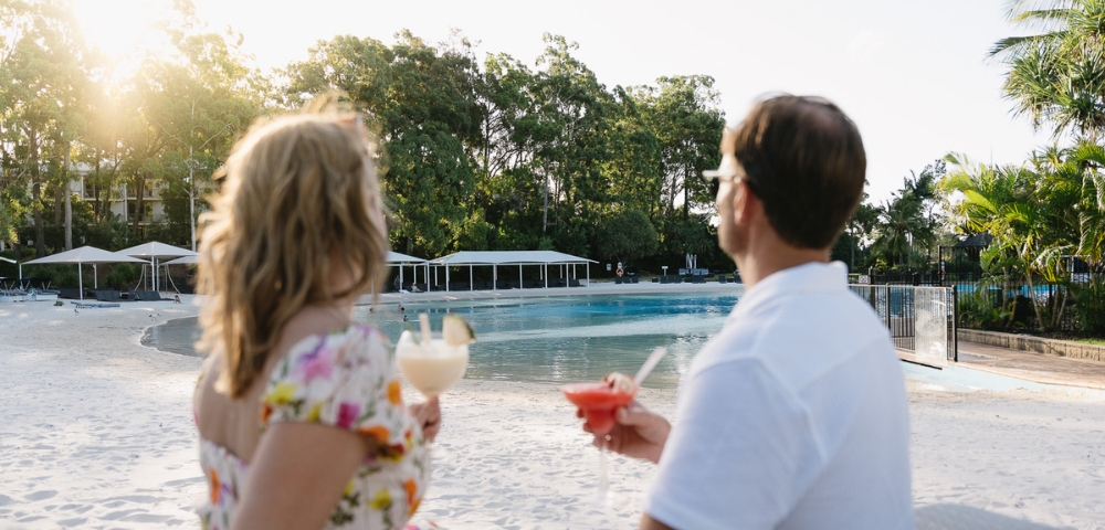 A couple enjoys tropical cocktails by a serene poolside at sunset, surrounded by lush greenery and white cabanas.