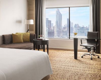 Modern hotel room with a bed, seating area, workspace, and large window showcasing a city skyline.