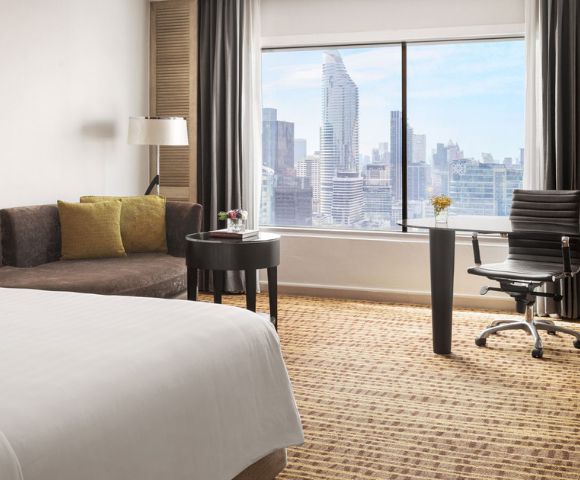 Modern hotel room with a bed, seating area, workspace, and large window showcasing a city skyline.