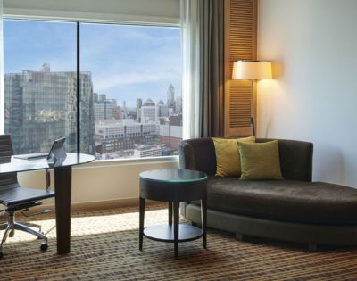 A modern hotel room featuring a cozy sofa, a round table, a desk with a chair, and a panoramic city view through large windows.