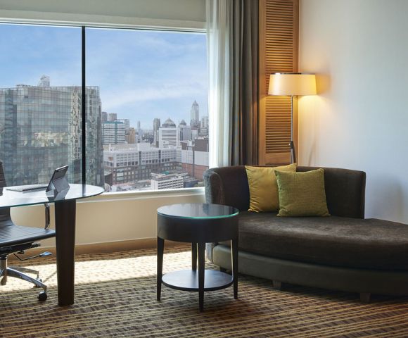 A modern hotel room featuring a cozy sofa, a round table, a desk with a chair, and a panoramic city view through large windows.