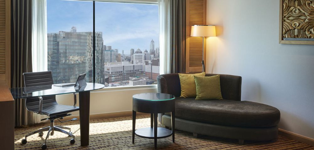 A modern hotel room featuring a cozy sofa, a round table, a desk with a chair, and a panoramic city view through large windows.