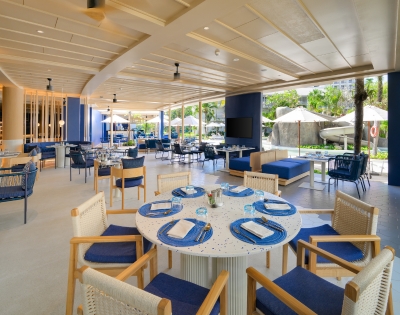 Bright, modern restaurant with blue accents, featuring round tables set for dining and large windows overlooking an outdoor seating area.