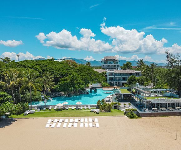 Aerial view of a beach resort with a large pool, palm trees, and lounge chairs under a bright blue sky with fluffy clouds.