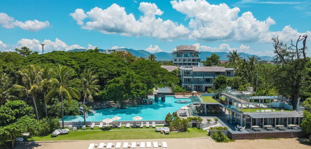 Aerial view of a beach resort with a large pool, palm trees, and lounge chairs under a bright blue sky with fluffy clouds.