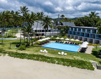 Aerial view of a tropical resort featuring a pool, sun loungers, palm trees, and modern accommodations against a cloudy sky.