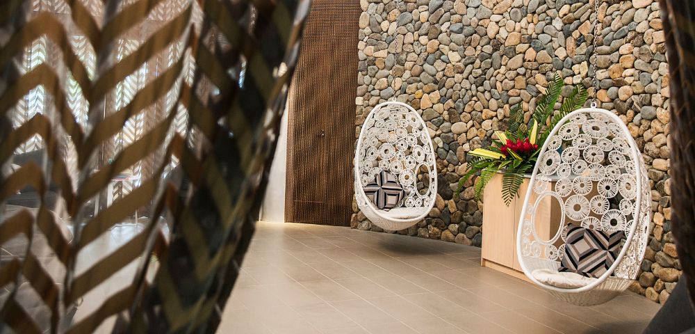 A cozy interior featuring a hanging white chair and a decorative stone wall, alongside a tropical plant arrangement.