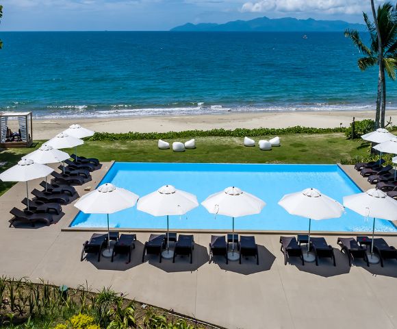 A serene beachfront view featuring a blue swimming pool surrounded by lounge chairs and umbrellas, framed by palm trees and ocean waves.