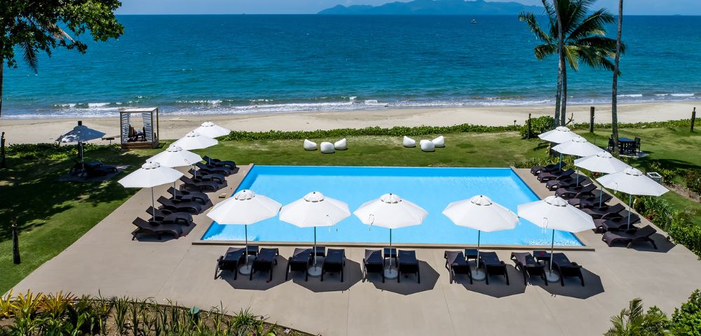 A serene beachfront view featuring a blue swimming pool surrounded by lounge chairs and umbrellas, framed by palm trees and ocean waves.