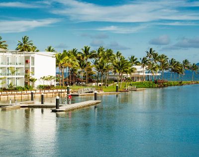 Tropical waterfront scene featuring palm trees, modern white buildings, and a calm blue bay with a wooden dock. Clear skies above.