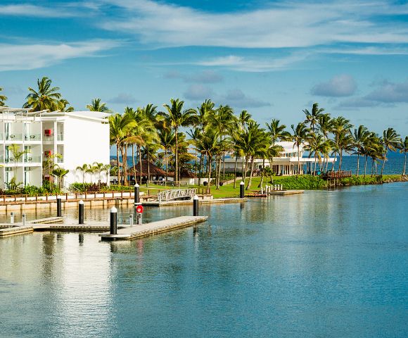 Tropical waterfront scene featuring palm trees, modern white buildings, and a calm blue bay with a wooden dock. Clear skies above.