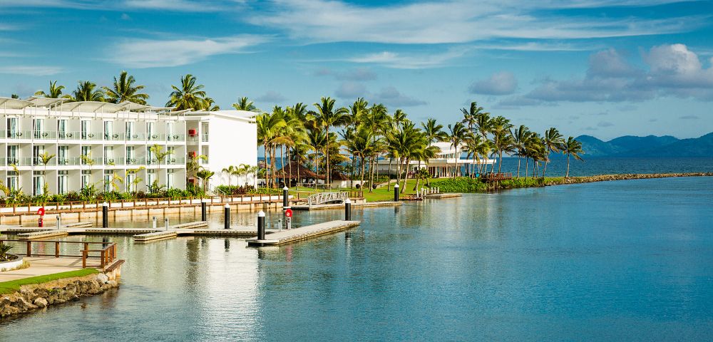 Tropical waterfront scene featuring palm trees, modern white buildings, and a calm blue bay with a wooden dock. Clear skies above.