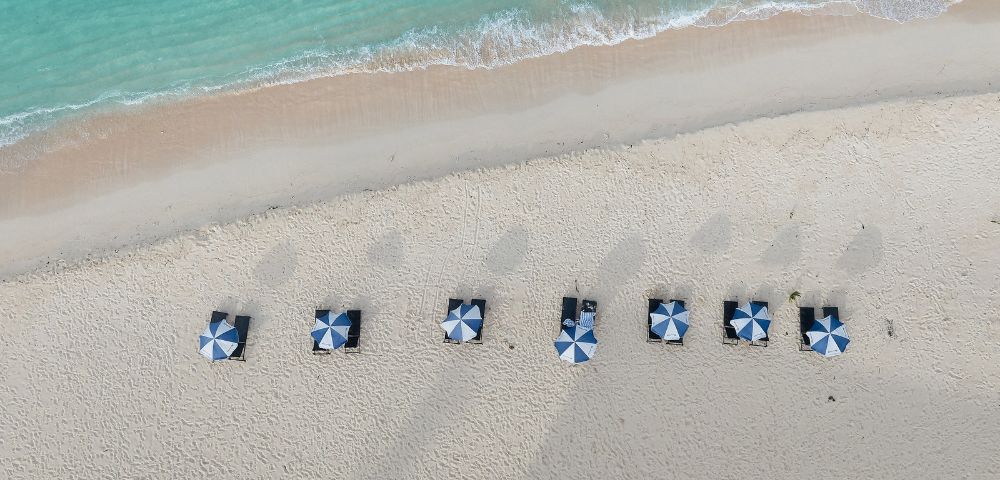 Aerial view of a sandy beach with striped blue and white umbrellas arranged in neat rows along the shoreline by turquoise waters.