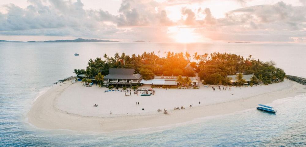 Aerial view of a tropical island during sunset, featuring palm trees, a sandy beach, and people enjoying the serene environment.