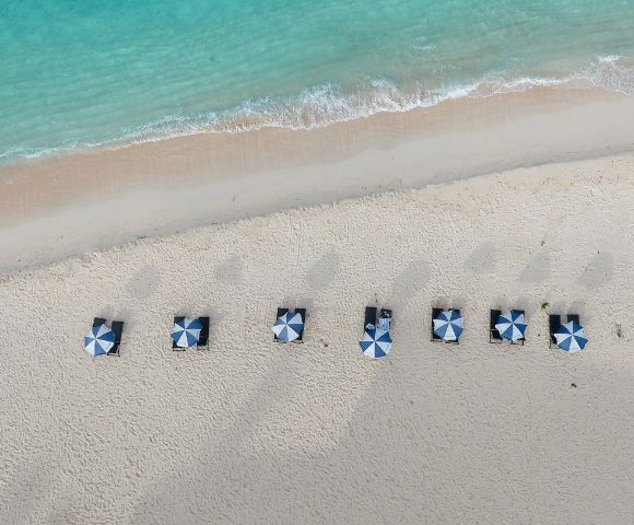 Aerial view of a sandy beach with striped blue and white umbrellas arranged in neat rows along the shoreline by turquoise waters.
