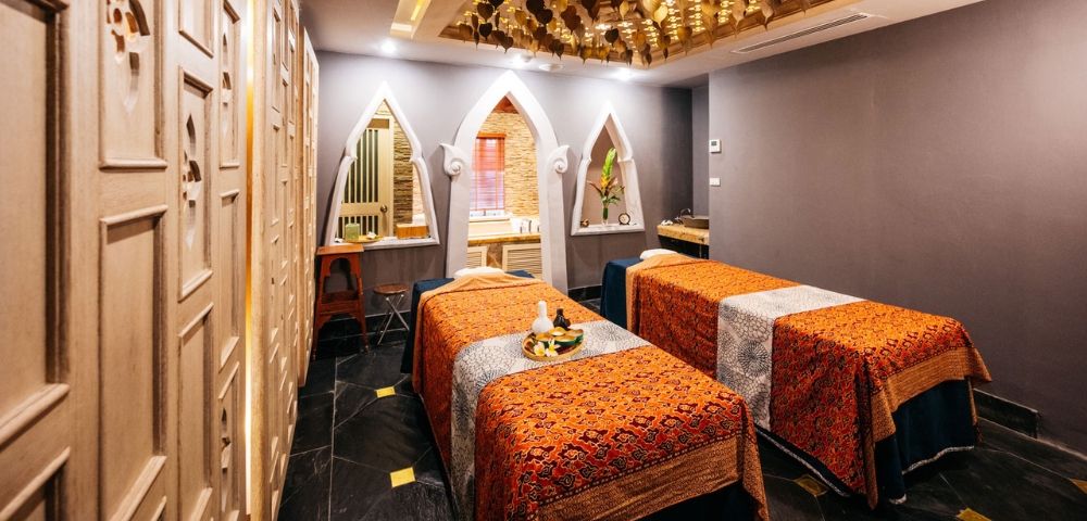 A serene spa room featuring two massage tables adorned with colorful fabric, decorative lighting, and elegant architecture.