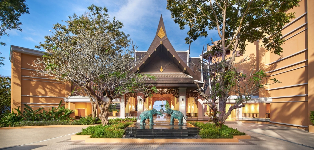 Entrance to a luxurious hotel featuring traditional architecture, lush trees, and bronze elephant sculptures amid a clear blue sky.
