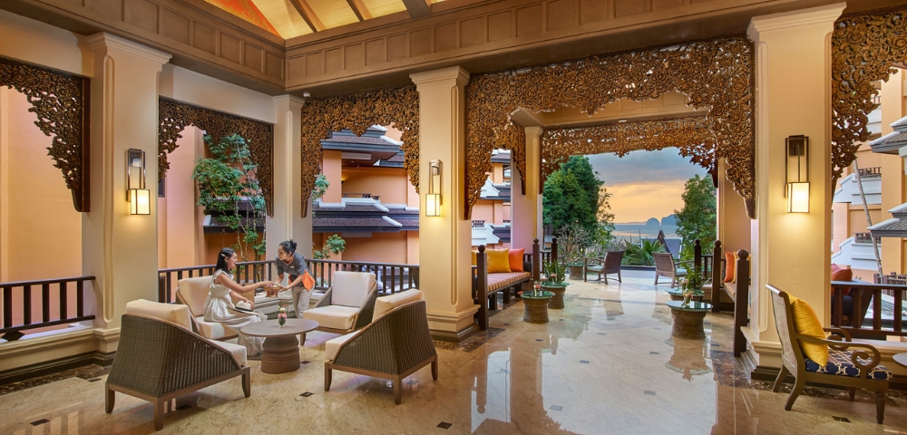 Two people converse in a luxurious, open-air lounge with elegant decor, palm trees, and a sunset view.