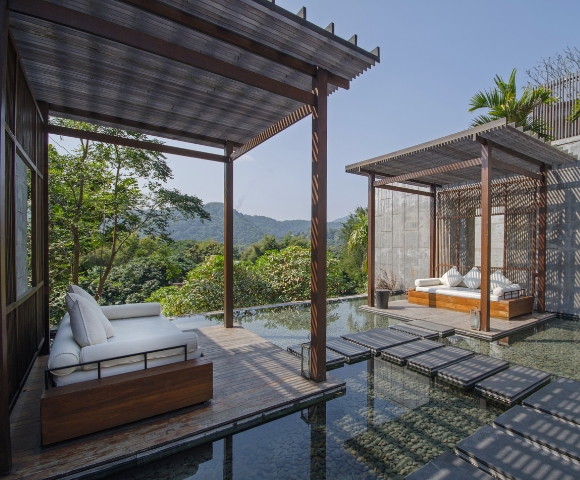 Modern outdoor lounge area with wooden pavilions overlooking lush greenery and mountains, featuring cozy seating and glassy water surface.
