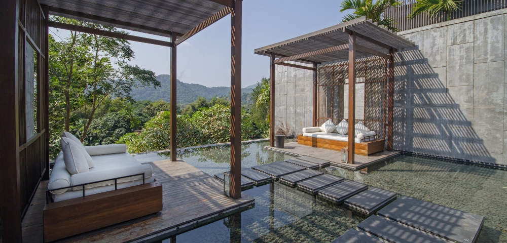 Modern outdoor lounge area with wooden pavilions overlooking lush greenery and mountains, featuring cozy seating and glassy water surface.