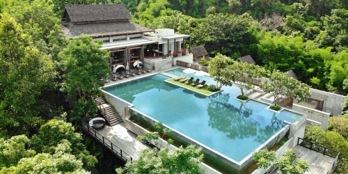 Aerial view of a luxurious resort featuring a large pool surrounded by lush greenery and a modern building in the background.